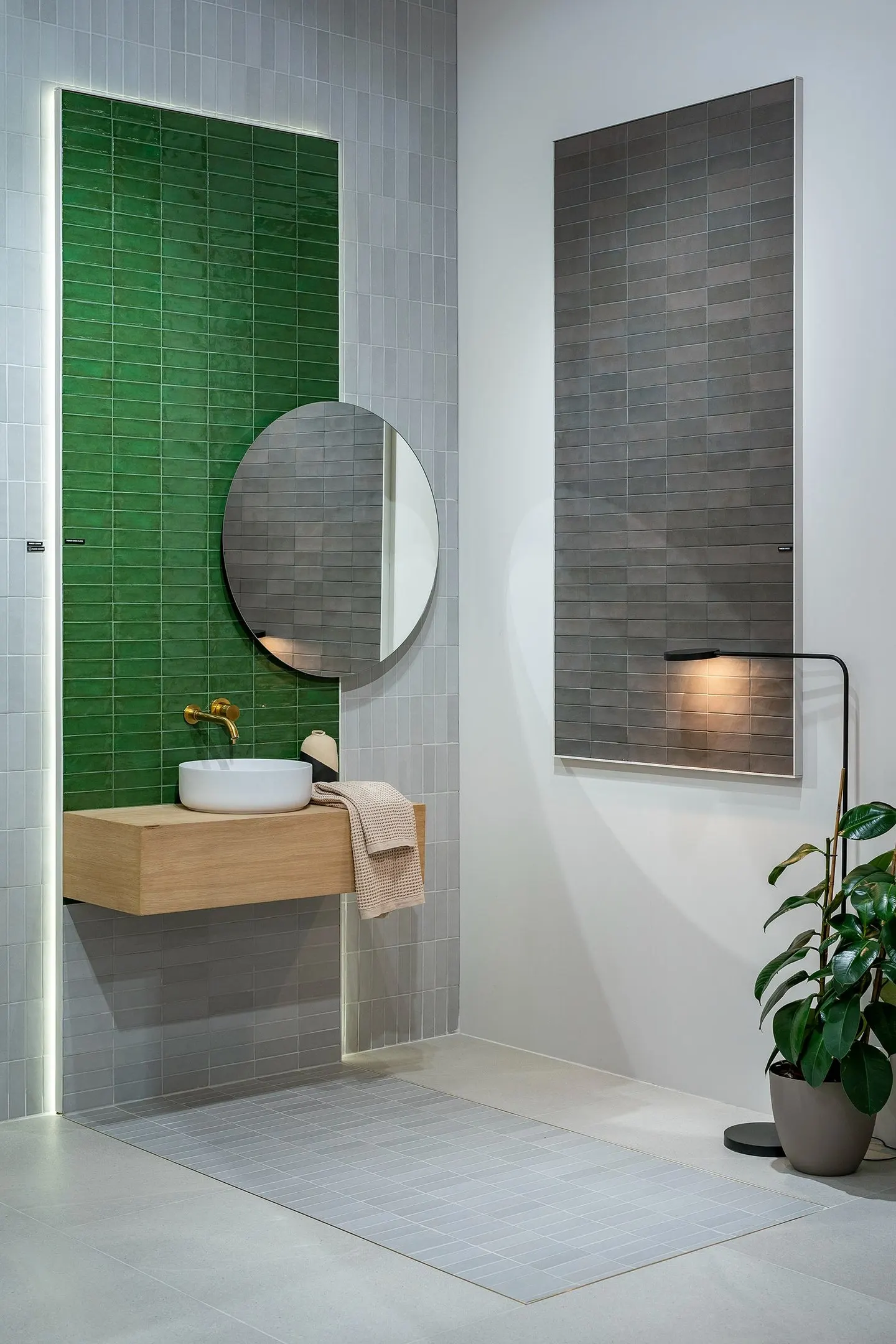 Baño de exposición con pared verde alicatada y espejo redondo